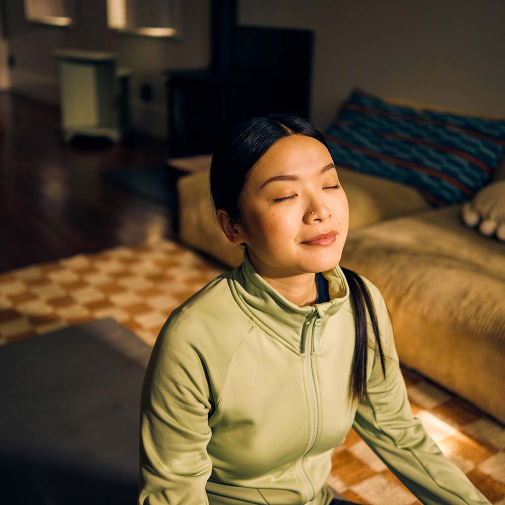 woman sitting on a yoga mat, with a slight smile on her face as she closes her eyes and faces the sun