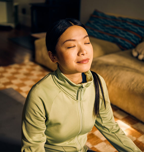 woman sitting on a yoga mat, with a slight smile on her face as she closes her eyes and faces the sun