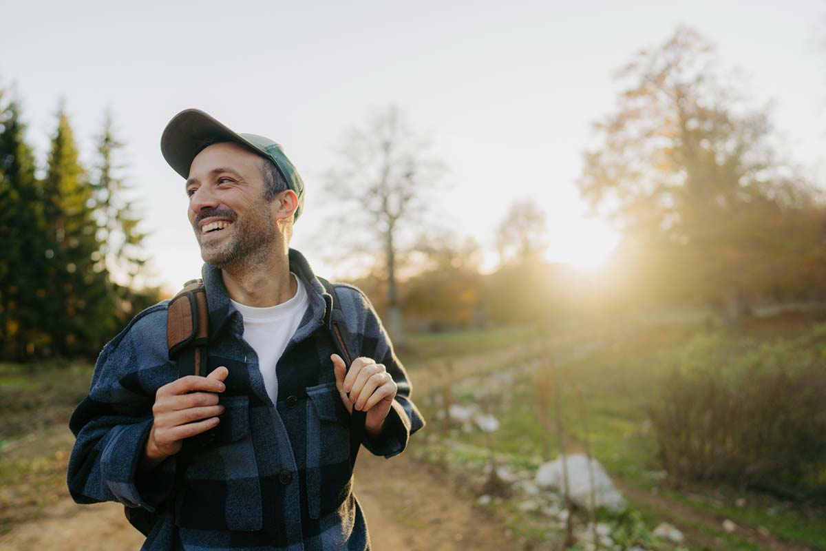 a man in a flannel shirt and a backpack smiles and hikes down the trail