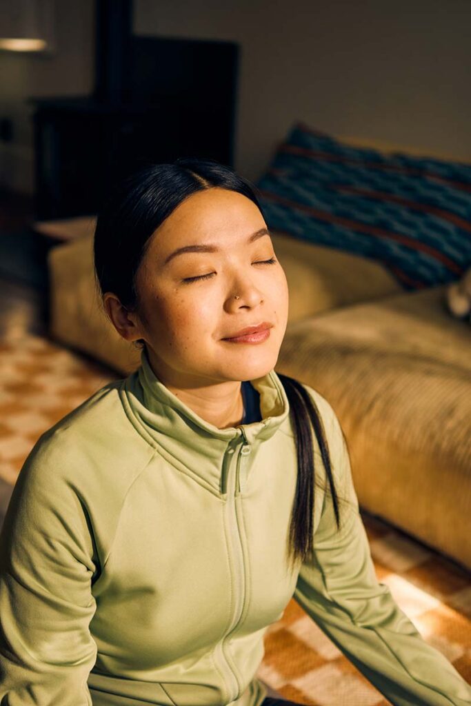 woman sitting on a yoga mat, with a slight smile on her face as she closes her eyes and faces the sun