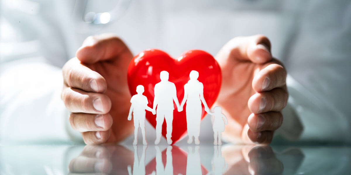 A doctor's hands cup a red heart shielding paper-cutout figures of a family holding hands.