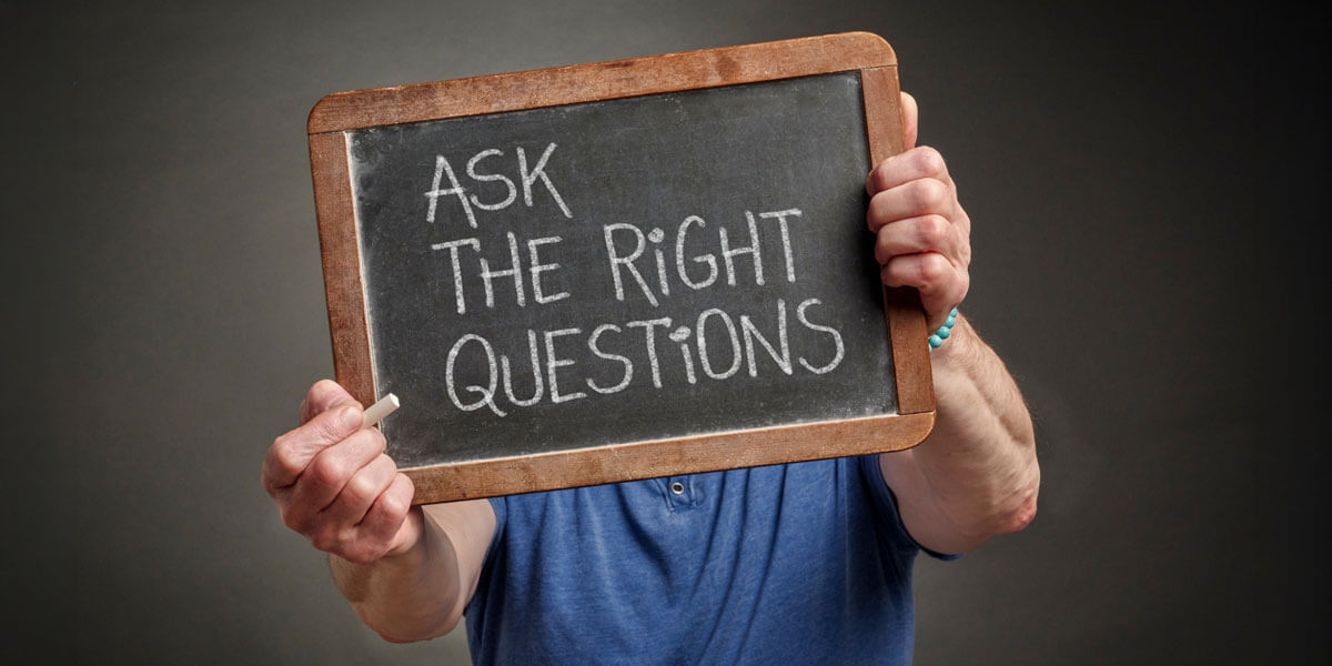 A person holds a small chalkboard reading "Ask the right questions" in front of their face.