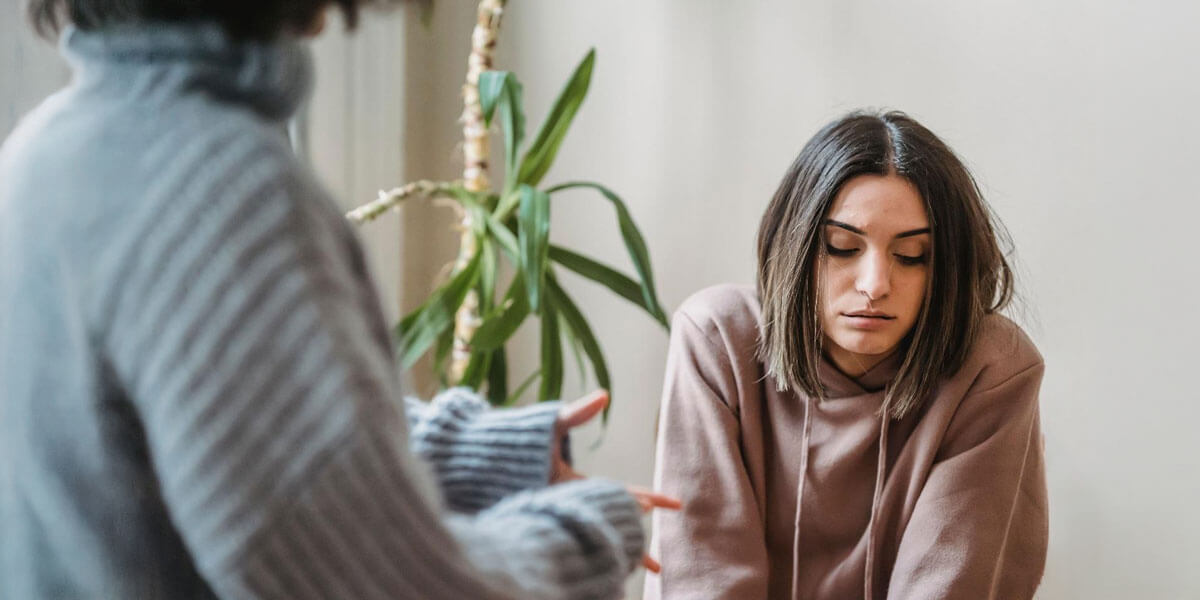 A woman sits looking down while another person gestures with hands nearby, suggesting a supportive conversation.