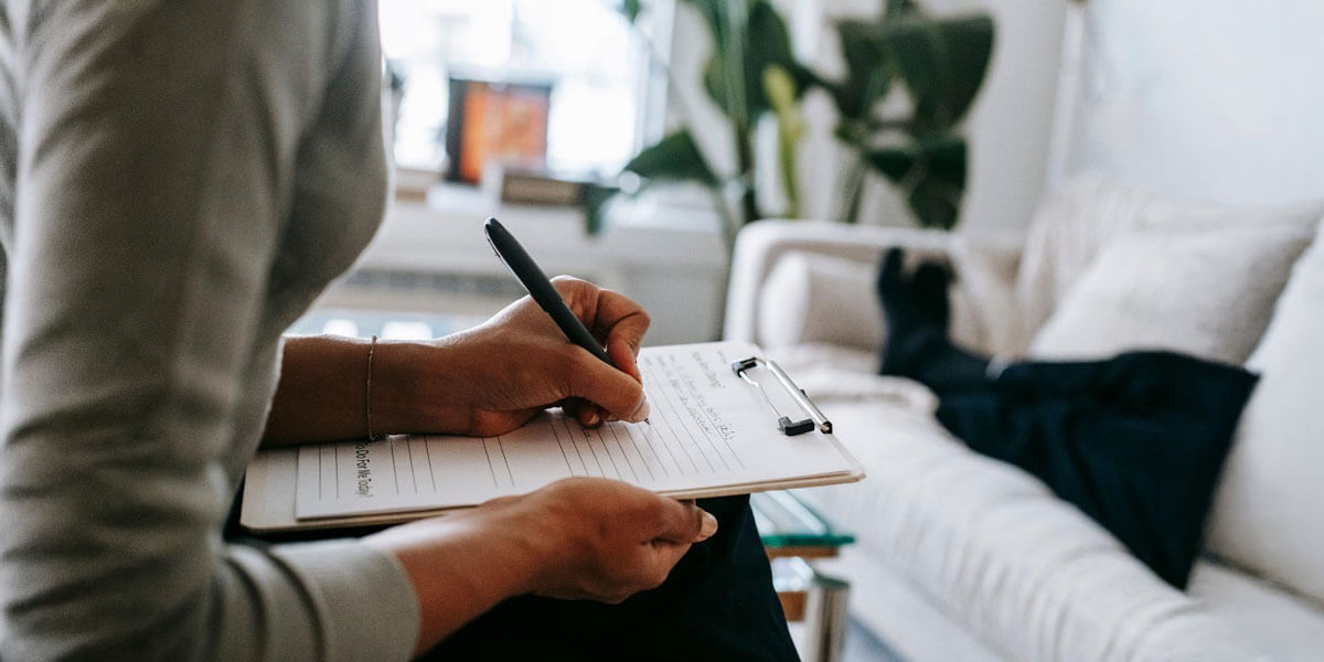 A close-up of a person writing notes on a clipboard in a bright, plant-filled living room.