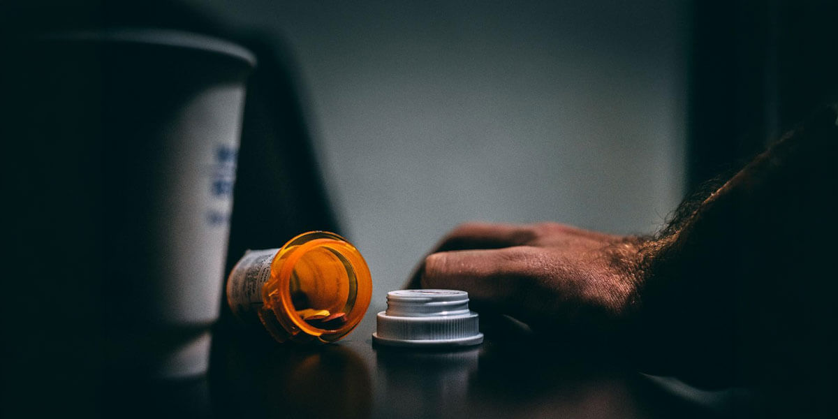 Empty pill bottle tipped over on a dark table beside a cup near a man's resting hand.