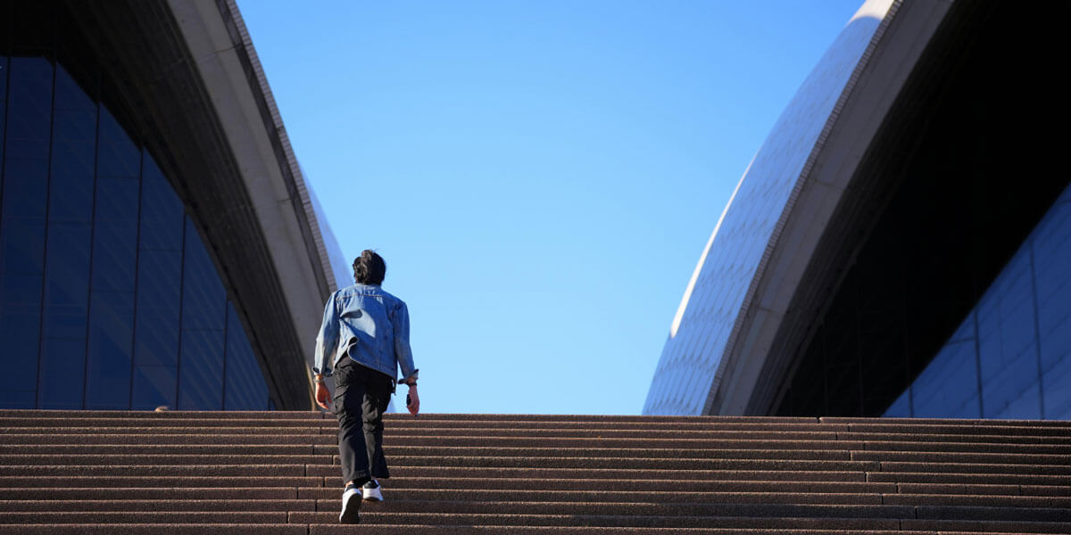 A person in a denim jacket climbs a long staircase toward a clear blue sky framed by the curved white sails of a large modern building.