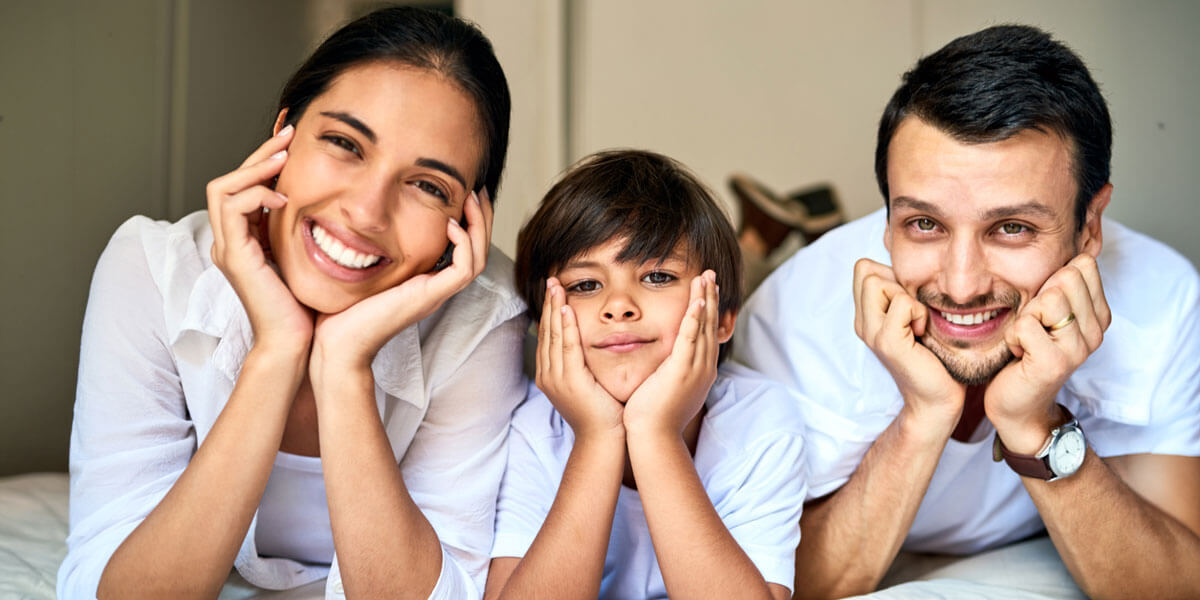 A smiling mother, son, and father lie side by side propping their chins on their hands, posing happily together on a bed.