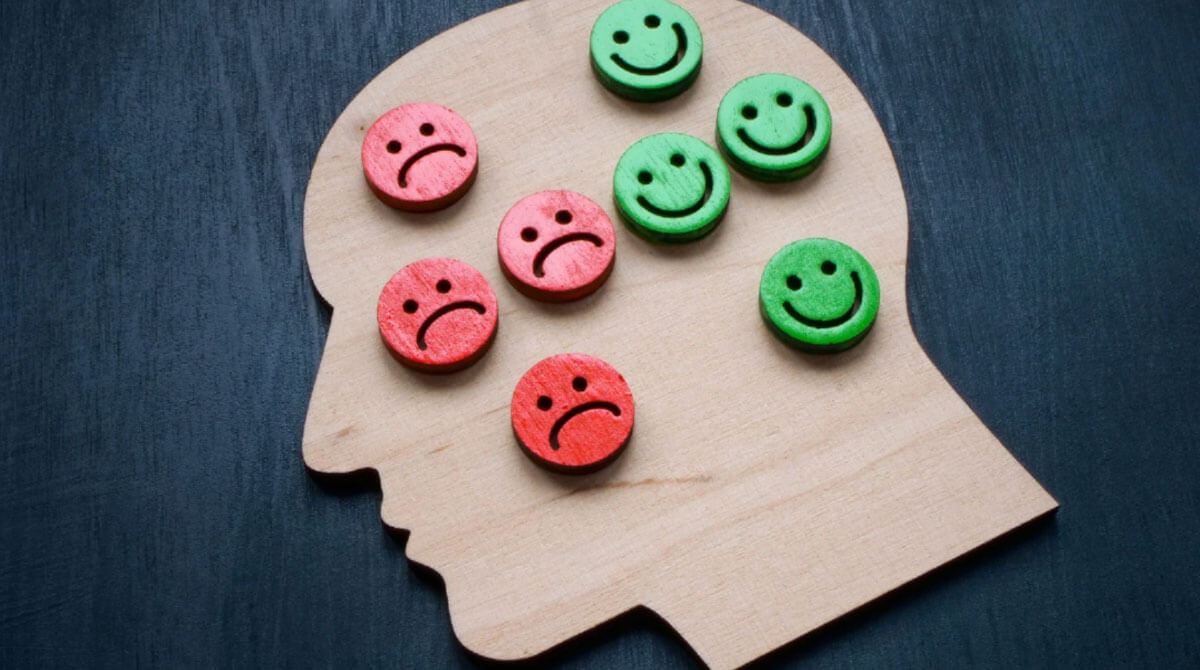 Wooden head silhouette filled with red sad and green happy face icons, representing mental health and emotional balance.