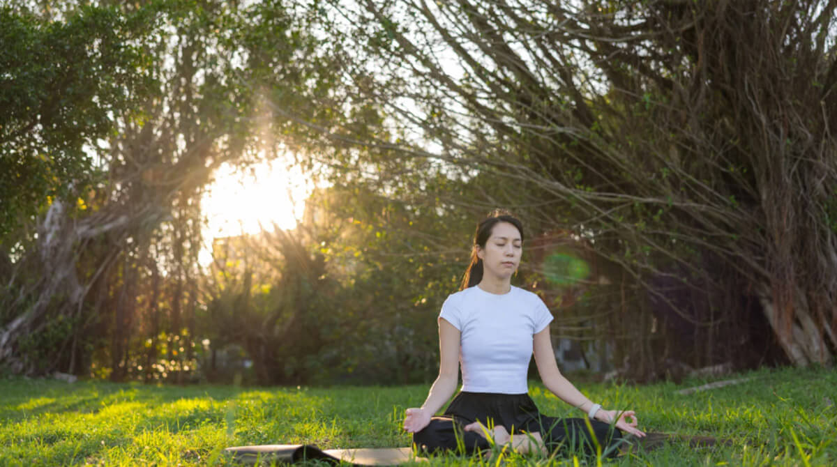 A young woman sits cross-legged on grass outdoors, meditating peacefully in a natural setting with sunlight filtering through trees.