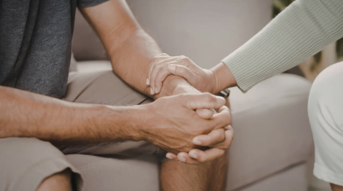 A close-up shows one person placing a reassuring hand over another’s hands, expressing comfort and emotional support.