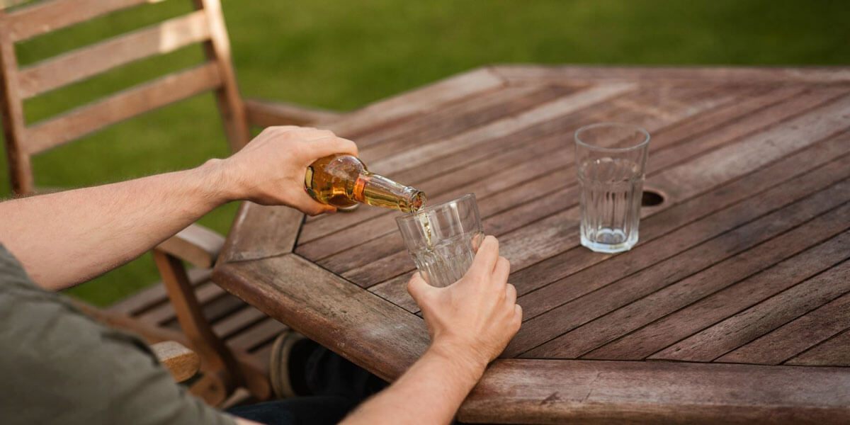 Man pouring a bottle of beer into a glass while seated at an outdoor wooden table.