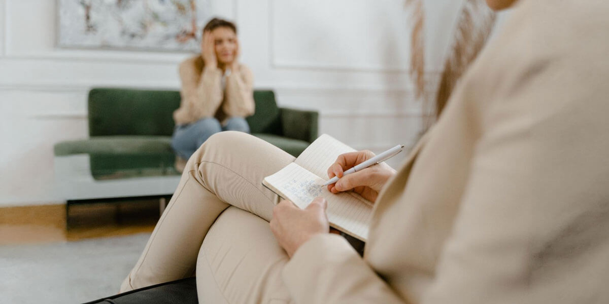 Distressed woman on a green couch being observed by a therapist writing notes.